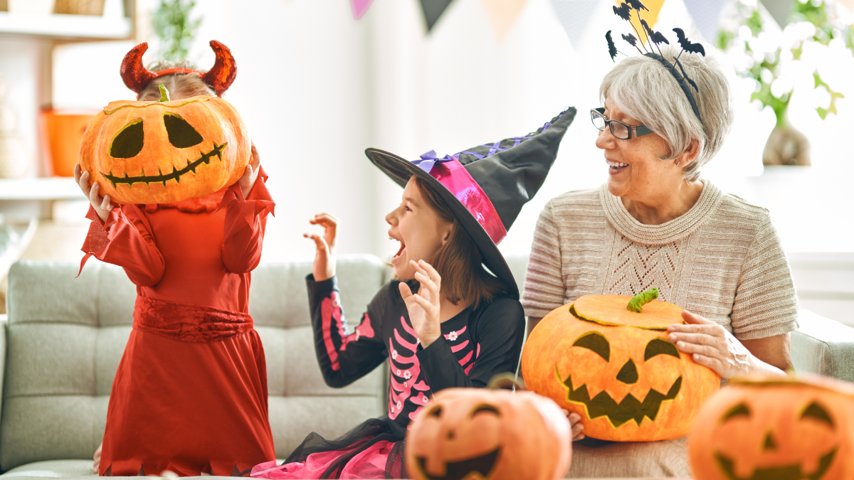 Kids in Halloween costumes and a grandmother holding jack-o’-lantern pumpkins, laughing and enjoying festive decorations at home.